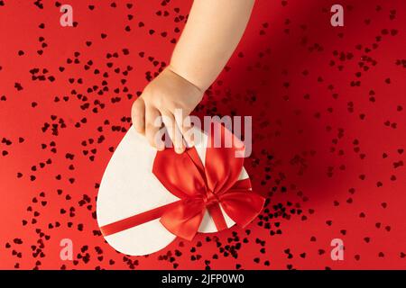 Foto von Valentinstag Dekoration von oben weiße herzförmige Geschenkbox mit roten Satinband Schleife und Konfetti auf rotem Hintergrund.Eine Grußkarte. Stockfoto