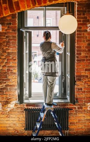 Eine Arbeiterin wäscht das Fenster, während sie mit dem Rücken zu uns auf einer Stehleiter steht Stockfoto