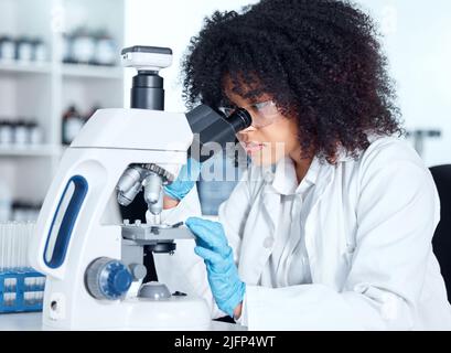 Eine junge afroamerikanische Frau, die einen Labormantel und eine Brille trägt und medizinische Proben auf einem Mikroskop in ihrem Labor ansieht. Eine Wissenschaftlerin mit gemischter Rasse Stockfoto