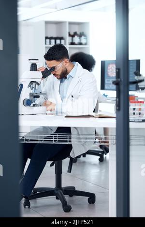 Ein gemischter Rennwissenschaftler, der eine Schutzbrille trägt, während er medizinische Testproben mit einem Mikroskop in einem Labor analysiert. Junger indischer Mann ermittelt mit Stockfoto