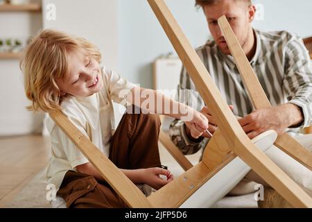 Porträt von niedlichen blonden Jungen helfen Vater Montage von Holzmöbeln zu Hause in erdigen gemütlichen Tönen Stockfoto