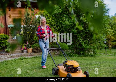 Ältere Frau mäht Gras mit Rasenmäher im Garten, Gartenarbeit Konzept. Stockfoto