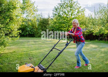 Ältere Frau mäht Gras mit Rasenmäher im Garten, Gartenarbeit Konzept. Stockfoto