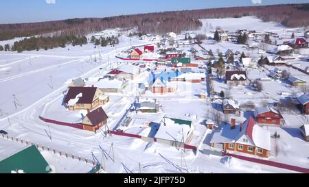 Der Blick von der Drohne. Clip.Winterwälder, wo kleine Häuser des Dorfes sichtbar sind und große Schneeverwehungen und blauer Himmel und große lange grüne Stockfoto