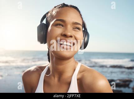 Junge, gemischte Sportlerin, die glücklich und lächelnd aussieht, während sie Kopfhörer trägt und Musik hört, während sie auf der Promenade läuft Stockfoto