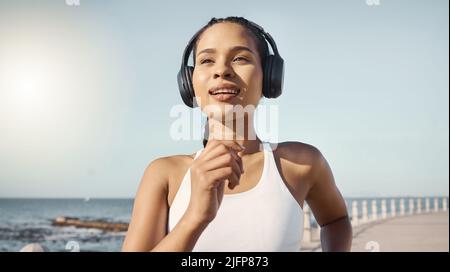 Fröhliche junge Fitnessfrau in Sportkleidung, die kabellose Kopfhörer trägt und Musik hört, während sie auf der Promenade läuft. Bewegung ist gut Stockfoto