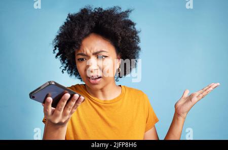 Dieser Text macht keinen Sinn. Aufnahme einer jungen Frau, die im Studio vor blauem Hintergrund steht und verwirrt aussieht, während sie ihr Handy benutzt. Stockfoto