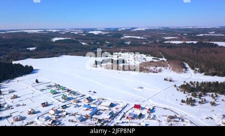 Der Blick von der Drohne. Clip.Winterwälder, wo kleine Häuser des Dorfes sichtbar sind und große Schneeverwehungen und blauer Himmel und große lange grüne Stockfoto