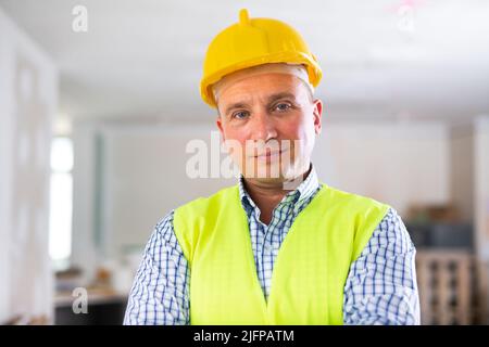 Porträt eines lächelnden Arbeiters in gelber Weste und Hut in der Hütte, die gerade renoviert wird Stockfoto