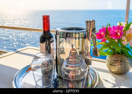 Eine Flasche Rotwein mit Blick auf den Sonnenuntergang, ein Eiskübel und zwei stemless Gläser neben einem Blumenstrauß auf dem Balkon einer Kreuzfahrt-Kabine auf dem Meer. Stockfoto