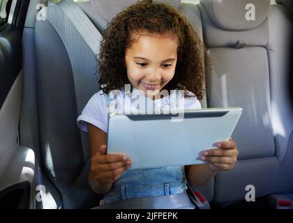 Unterhaltung an Bord. Ein kurzer Schuss eines entzückenden kleinen Mädchens, das ihr Tablet benutzt, während es auf dem Rücksitz eines Autos sitzt. Stockfoto