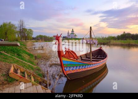 Boot auf dem Fluss Kamenka. Tourist drakkar in der Nähe des Ufers an der Iljinsky Kirche. Susdal, Russland, 2022 Stockfoto