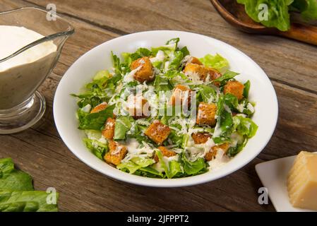 Caesar-Salat in einer weißen Schüssel auf einem alten rustikalen Holztisch mit Blättern mit einem Saucenboot und Parmesan. Ausgewählter Fokus. Stockfoto