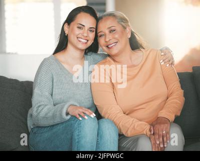 Teil einer Familie zu sein bedeutet, für Fotos zu lächeln. Aufnahme einer Mutter und Tochter, die zu Hause auf dem Sofa sitzen. Stockfoto