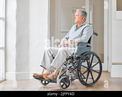 Ich wünschte, ich wäre in einer besseren Phase meines Lebens. Aufnahme eines älteren Mannes, der in einem Rollstuhl sitzt und zu Hause nachdenklich aus dem Fenster schaut. Stockfoto