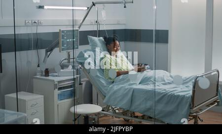 Kranker Patient, der sein Mobiltelefon hält, um mit Freunden über Videoanruf zu sprechen. Frau mit Krankheit, die Online-Videokonferenzen nutzt, um mit der Familie auf der Krankenhausstation zu sprechen. Stockfoto