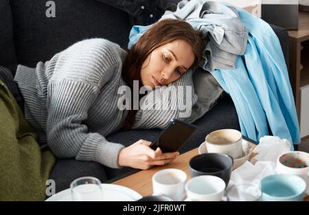 Wenn es um Besessenheit geht. Aufnahme einer jungen Frau, die auf ihrer unordentlichen Couch liegt und deprimiert aussieht und ihr Smartphone benutzt. Stockfoto