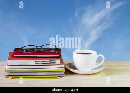 Kaffeetasse, Gläser und Bücherstapel auf einem Holztisch mit unscharfem Himmel. Geschäftskonzept Stockfoto