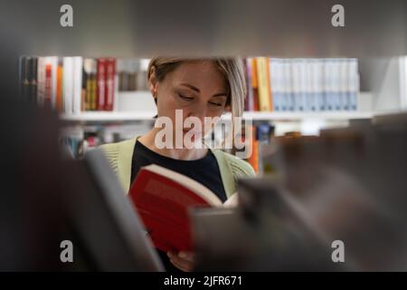 Reife Studentin, die im Bücherregal der Bibliothek nach Büchern sucht, die auf dem Universitätscampus oder College studieren Stockfoto