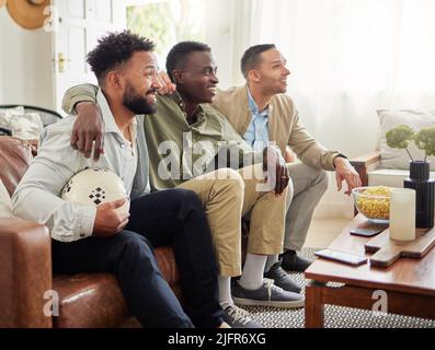 Es war ein Spieltag und wir mussten uns einfach nur treffen. Aufnahme von drei männlichen Freunden, die zusammen etwas beobachten, während sie auf einer Couch sitzen. Stockfoto