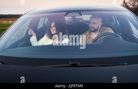 Vorderansicht im schwarzen Auto eines fokussierten männlichen Fahrers und einer lächelnden Beifahrerin, die mit dem Finger auf die Landschaft auf der linken Seite zeigt. Zwei Personen, die mit dem Auto in die Stadt fahren. Stockfoto