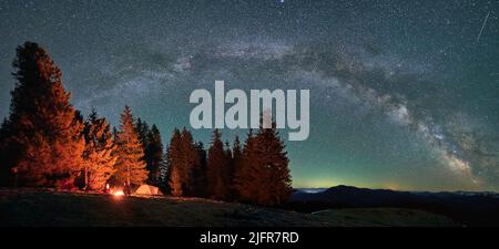 Panorama des Sternenhimmels, Milchstraße über die Berge. Silhouetten von zwei Reisenden mit Zelt, die sich durch Lagerfeuer erwärmen. Wald, Gipfel von Bergen mit Licht von Siedlungen in der Ferne im Hintergrund. Stockfoto