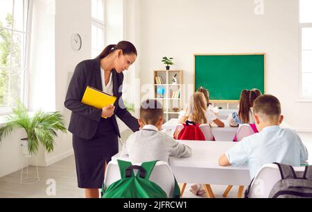 Die Lehrerin hilft ihren Grundschülerinnen, die an Schreibtischen sitzen und schreiben Stockfoto