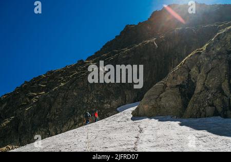 Kletter-Team Ropping up Frau und Mann gekleidet Höhenbergsteigen Kleidung mit Rucksäcken zu Fuß durch verschneiten Hang in der felsigen Couloir. Aktiv Stockfoto