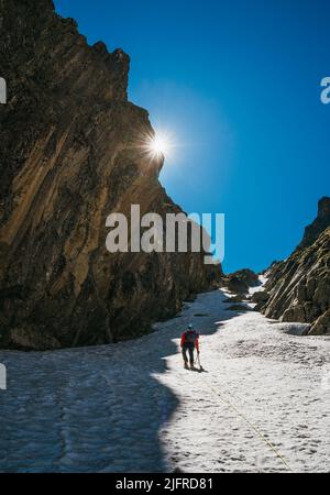 Team Roping up Frau mit Kletteraxt gekleidet Höhenbergsteigen Kleidung mit Rucksack zu Fuß durch verschneiten Hang im Couloir mit Hintergrundbeleuchtung Stockfoto