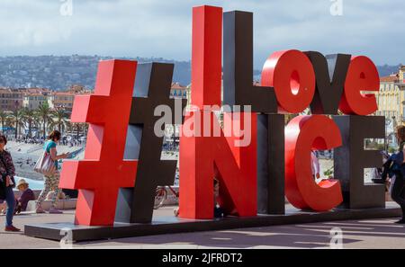 Nizza, Frankreich, April 2022. Rot-schwarzes Touristenschild „I love Nice“ über der englischen Promenade. Touristen fotografieren am Schild. Stockfoto