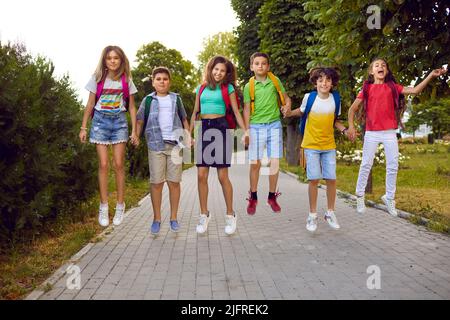 Lustige Grundschüler mit Rucksäcken auf den Schultern haben Spaß, im Park zusammen zu springen Stockfoto