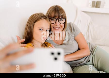 Das weiße Mädchen und ihre Großmutter machen Selfie auf dem Handy, während sie zu Hause auf dem Sofa sitzen Stockfoto