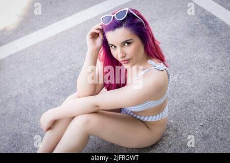 Seitenansicht einer jungen Frau in einer stilvollen Sonnenbrille, die Haare berührt und die Kamera anschaut, während sie auf einer Asphaltstraße auf der Stadtstraße sitzt Stockfoto