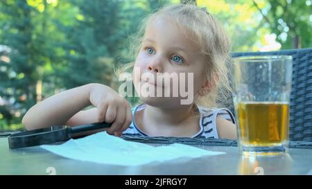 Odessa, Ukraine, Osteuropa. 4.. Juli 2022. Nettes kleines Mädchen sitzt nachdenklich am Tisch mit einer Linse in der Hand. Nahaufnahme eines blonden Mädchens, das eine Lupe in der Hand hält und an etwas denkt, während es im Straßencafé im Park sitzt. (Bild: © Andrey Nekrasov/ZUMA Press Wire) Stockfoto