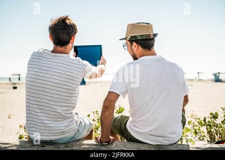 Porträt eines jungen Mannes, der bei Sonnenaufgang mit einem Tablet am Sandstrand sitzt. Reise-, Personen-, Urlaub- und Technologiekonzept Stockfoto