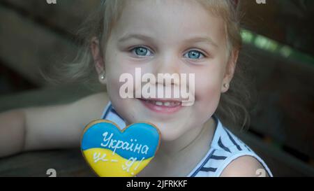 Das kleine blonde Mädchen hält Lebkuchen in ukrainischen Nationalfarben in der Hand und sagt: „Die Ukraine ist ich“. Nahaufnahme Porträt des Mädchens auf Park benc sitzen Stockfoto