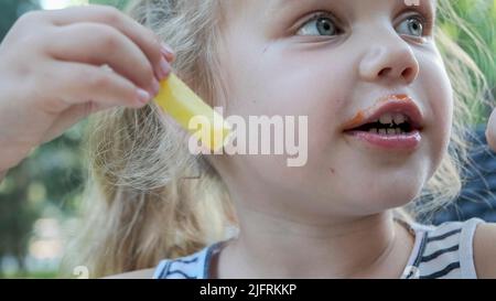 Kleine Mädchen essen pommes. Nahaufnahme des blonden Mädchens nimmt Kartoffelchips mit ihren Händen und probiert sie im Straßencafé im Park. Stockfoto