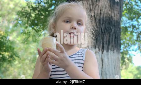 Nettes kleines Mädchen isst draußen Eis. Nahaufnahme eines blonden Mädchens, das auf der Parkbank sitzt und Eis isst. Stockfoto