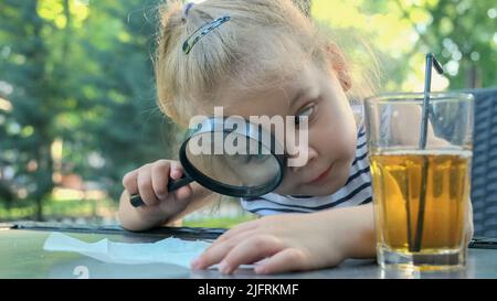 Kleines Mädchen schaut vorsichtig in die Linse auf das Salz. Nahaufnahme des blonden Mädchens studiert Salzkristalle, während sie durch die Lupe betrachtet wird Stockfoto