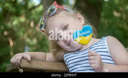 Odessa, Ukraine, Osteuropa. 3.. Juli 2022. Das kleine blonde Mädchen hält Lebkuchen in ukrainischen Nationalfarben in der Hand und sagt: „Die Ukraine ist ich“. Nahaufnahme des Mädchens, das auf der Parkbank sitzt, mit Cookies in ukrainischen Flaggen-Farben. Zeitlupe. Odessa Ukraine (Foto: © Andrey Nekrasov/ZUMA Press Wire) Stockfoto