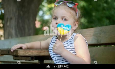 Odessa, Ukraine, Osteuropa. 4.. Juli 2022. Das kleine blonde Mädchen hält Lebkuchen in ukrainischen Nationalfarben in der Hand und sagt: „Die Ukraine ist ich“. Nahaufnahme des Mädchens, das auf der Parkbank sitzt, mit Cookies in ukrainischen Flaggen-Farben. Zeitlupe. Odessa Ukraine (Foto: © Andrey Nekrasov/ZUMA Press Wire) Stockfoto