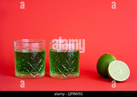 Grünes Alkoholgetränk in facettierten Gläsern mit Wassertropfen in der Nähe von Limetten auf Rot Stockfoto