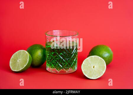 Grüner Alkoholdrink in facettiertem Glas in der Nähe von sauren Limetten auf rot Stockfoto