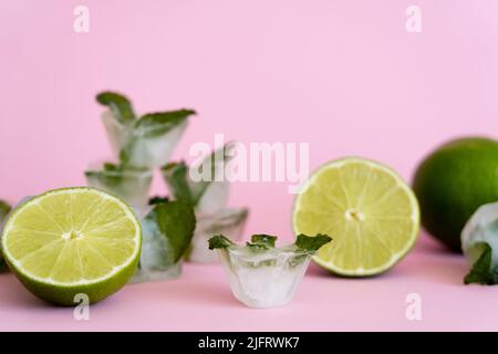 Grüne und frische Limetten in der Nähe verschwommener Eiswürfel mit Pfefferminze auf Rosa Stockfoto
