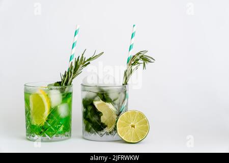 Kühle facettierte Gläser mit Cocktails und Eiswürfeln in der Nähe von Zitrusfrüchten auf Weiß