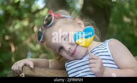 Odessa, Ukraine, Osteuropa. 3.. Juli 2022. Das kleine blonde Mädchen hält Lebkuchen in ukrainischen Nationalfarben in der Hand und sagt: „Die Ukraine ist ich“. Nahaufnahme des Mädchens, das auf der Parkbank sitzt, mit Cookies in ukrainischen Flaggen-Farben. Zeitlupe. Odessa Ukraine (Foto: © Andrey Nekrasov/ZUMA Press Wire) Stockfoto