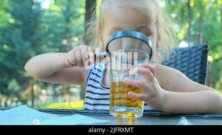 Odessa, Ukraine, Osteuropa. 4.. Juli 2022. Nettes kleines Mädchen schaut vorsichtig in die Linse auf den Saft im Glas. Nahaufnahme eines blonden Mädchens, das ihren Saft im Glas studiert, während es sie durch die Lupe betrachtet, während es im Straßencafé im Park sitzt. (Bild: © Andrey Nekrasov/ZUMA Press Wire) Stockfoto