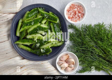 Zertrümmert leicht gesalzene gebrochene Gurken in einer Keramikschale mit rosa Salz, Knoblauch, Dill und Küchenhandtuchserviette. Draufsicht mit Kopierbereich Stockfoto