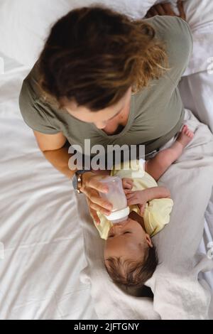 Direkt über der Aufnahme einer kaukasischen Mutter mittleren Erwachsenen, die zu Hause auf dem Bett neugeborenes Baby Milch füttert. Raum kopieren, unverändert, Familie, Liebe, Zweisamkeit, Pflege Stockfoto