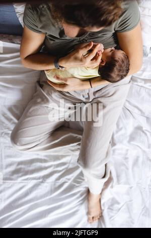 Direkt über der Aufnahme einer kaukasischen Mutter mit mittlerem Erwachsenen, die ein neugeborenes Baby stillt, während sie auf dem Bett sitzt. Raum kopieren, unverändert, Familie, Liebe, Zweisamkeit, Stockfoto